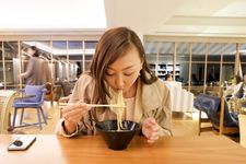 Seorang wanita memakai outfit blus dengan blazer sedang menyeruput ramen.