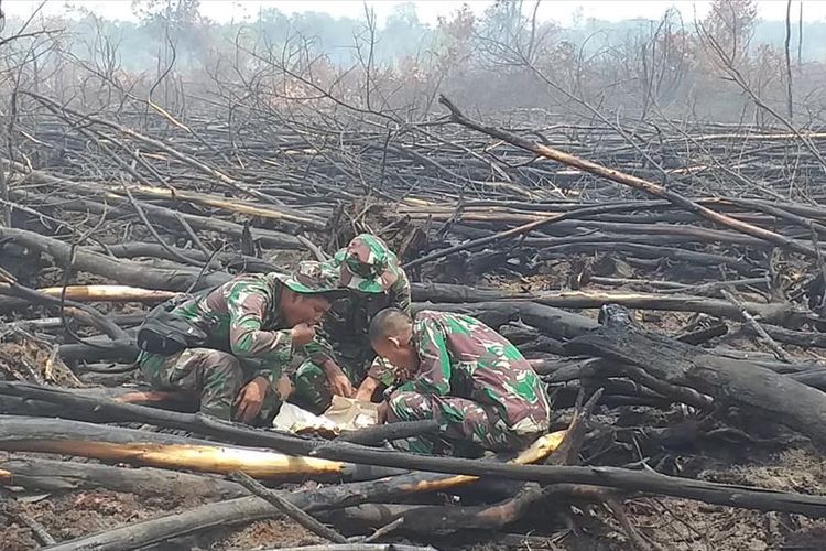 Serda Imam Effendi (tengah) dan dua prajurit TNI AD lainnya sedang makan siang di tumpukan kayu bekas terbakar di lahan yang sudah mereka padamkan di Desa Bedagu, Kecamatan Langgam, Kabupaten Pelalawan, Riau, Sabtu (3/8/2019).