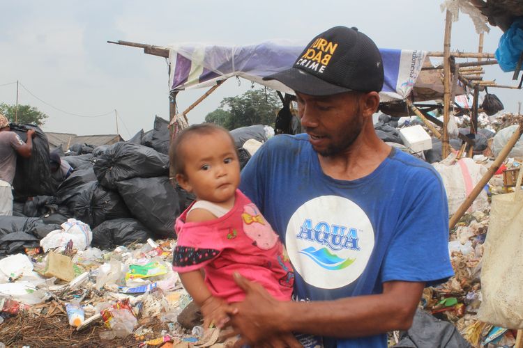 Tarudin (34) sedang menggendong anaknya di tengah lingkungan sekitar TPST Bantargebang. Mereka terlihat biasa saja menghirup udara dan bau dari tumpukan sampah tanpa alat pelindung apapun, termasuk masker.