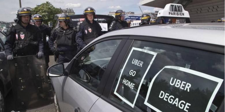 Polisi antihuru-hara berdiri di samping taksi yang ditempel poster yang berbunyi Uber pulang dan Uber keluar dalam demonstrasi di Paris, 25 Juni 2015. Sopir taksi di Paris melakukan aksi mogok dengan menutup jalan Bandara Charles de Gaulle.