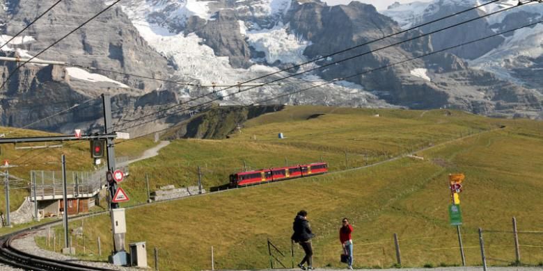 Wisatawan berpose dengan latar belakang kereta api listrik bergerigi (cogwheel) rute Jungfraujoch-Kleine Scheidegg yang tengah melaju di dinding Pegunungan Alpen, Swiss.