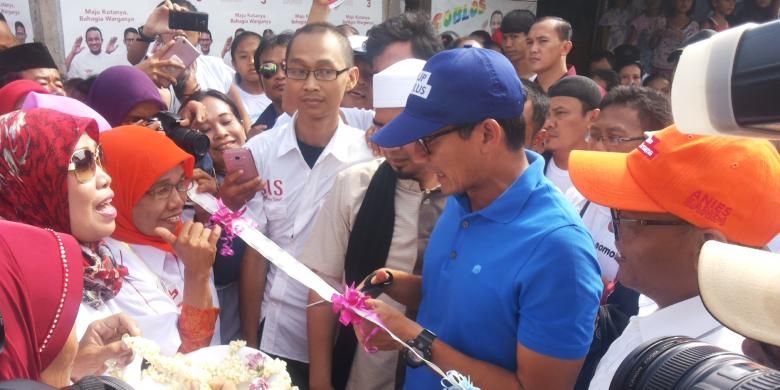 Calon wakil gubernur DKI Jakarta nomor pemilihan tiga Sandiaga Uno menggunting pita yang disiapkan warga saat kedatangannya di Kampung Kebon Bayam, Kelurahan Papanggo, Tanjung Priok, Jakarta Utara, Rabu (25/1/2017). Pita disiapkan warga untuk menandai perubahan nama kampungnya menjadi kampung Anies-Sandi.