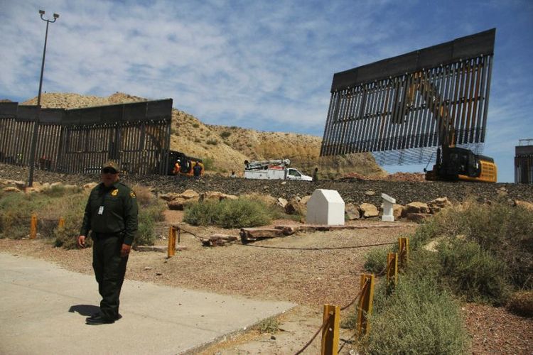 Seorang agen Patroli Perbatasan AS terlihat berada di sebelah para pekerja yang membangun pagar perbatasan di sebuah properti pribadi yang terletak di perbatasan Texas dan New Mexico dengan Ciudad Juarez, Meksiko, Minggu (26/5/2019). (AFP/HERIK MARTINEZ)
