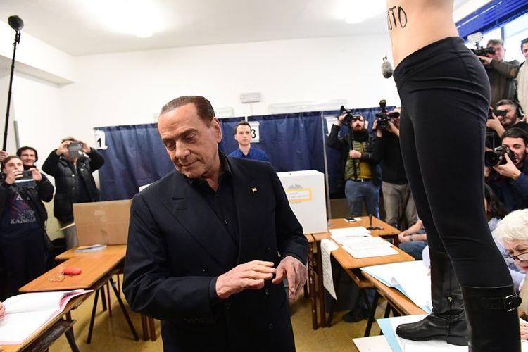 Mantan Perdana Menteri Italia, Silvio Berlusconi, langsung memalingkan mukanya ketika seorang perempuan berdiri di hadapannya sambil bertelanjang dada dengan tulisan BERLUSCONI SEI SCADUTO (Berlusconi, waktu Anda telah habis) ketika memberikan suara di Milan, Minggu (4/3/2018).