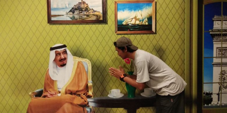 Pose Raja Salman terlihat sedang duduk termenung di kursi dengan dua buah gelas serta vas bunga terletak di atas meja. Ia seakan-akan memunculkan mimik muka terkejut melihat pengunjung yang datang.