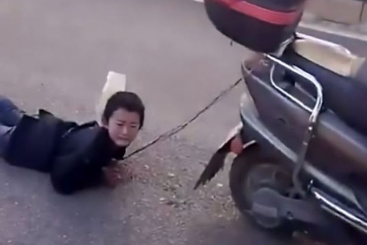 Dalam foto yang diambil dari video yang diunggah ke media sosial, terlihat seorang anak laki-laki yang menangis karena diseret ibunya dengan menggunakan sepeda motor di jalan raya.