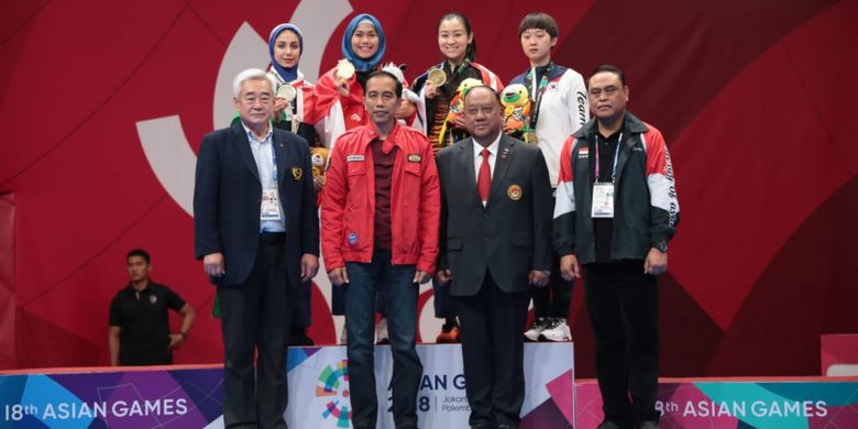 Defia Rosmaniar berfoto di belakang Presiden Republik Indonesia, Joko Widodo, bersama Chef de Mission (CdM) Tim Indonesia, Komjen Pol. Syafruddin, seusai pengalungan medali emas taekwondo di JCC Plenary Hall, 19 Agustus 2018.