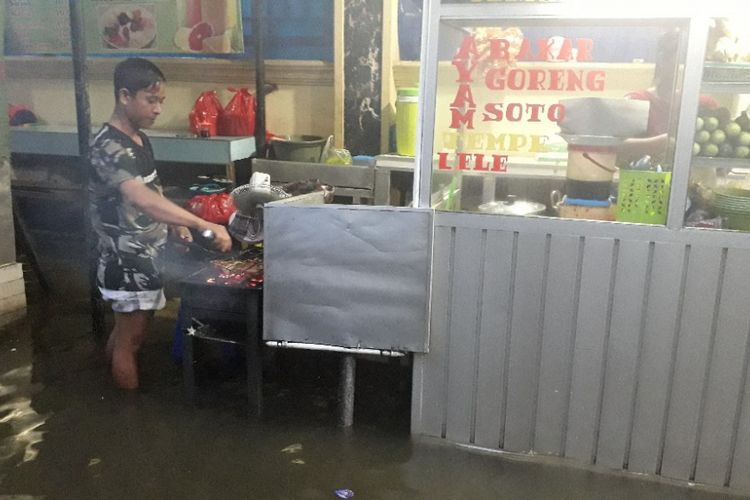 Seorang pedagang tengah memasak ayam bakar meski warungnya terendam banjir di Muara Angke, Jakarta Utara, Jumat (25/1/2019).