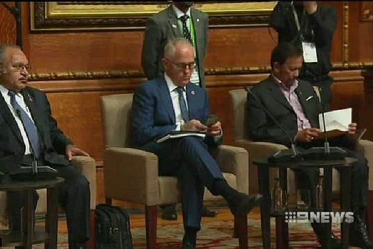 PM Malcolm Turnbull tertangkap kamera sedang sibuk memainkan ponselnya di saat PM Theresa May sedang berpidato di hadapan para pemimpin negara-negara Persemakmuran di London.