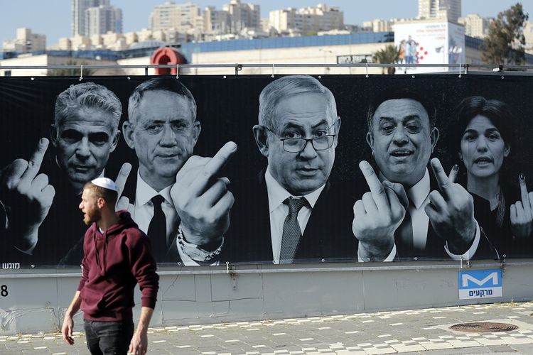Seorang warga Israel berjalan melewati sebuah poster kampanye pemilu yang menampilkan para politisi mengacungkan jari tengah, yang terpasang di Yerusalem.