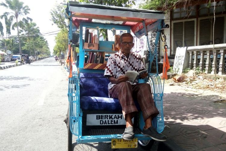 Sutopo saat membaca buka di becaknya sembari menunggu penumpang 