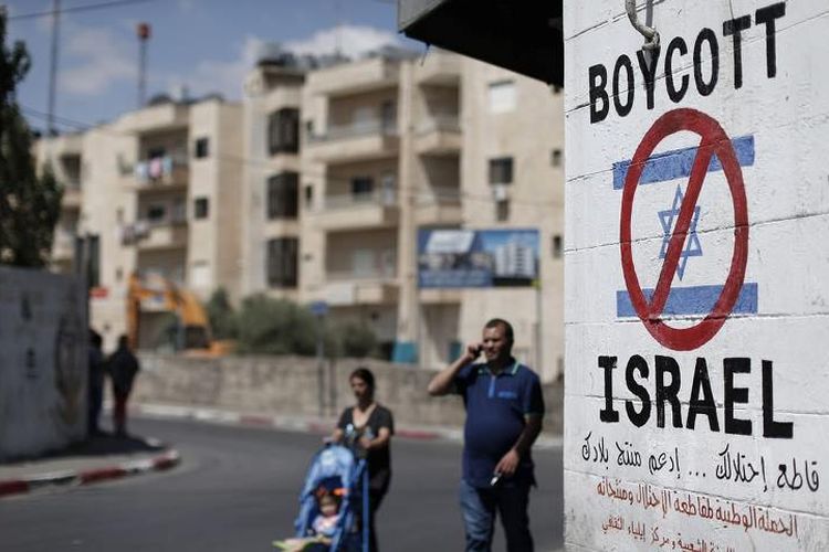 Warga Palestina berjalan di dekat sebuah tanda di tembok Bethlehem yang menyerukan untuk memboikot produk Israel.