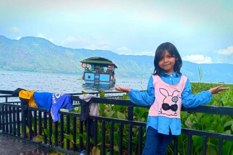 Seorang anak berfoto dengan latar belakang danau dan kapal di Dermaga Dedalu, Desa Dedalu, Kecamatan Lut Tawar, Kabupaten Aceh Tengah