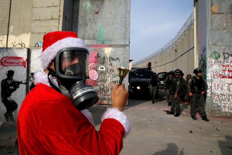 Warga Palestina mengenakan kostum Santa Claus, Sabtu (23/12/2017), berdiri di depan pasukan tentara Israel dengan memasang masker anti-gas air mata, di Betlehem, Yerusalem, sebagai bentuk protes terkait pernyataan Presiden Amerika Serikat Donald Trump yang mengakui Yerusalem sebagai ibu kota Israel. 
