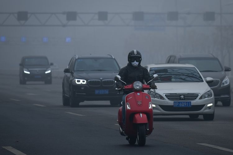 Pengendara motor mengenakan masker saat melintasi jalanan yang tertutup kabut asap tebal di Beijing, China.