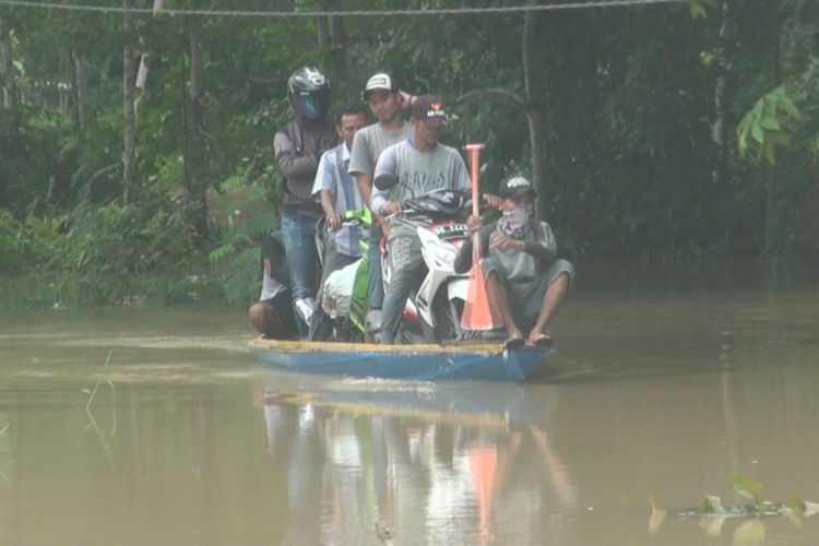 Sebuah perahu melintasi genangan banjir dengan membawa 2 unit sepeda motor beserta pemiliknya. Bnanir membuat warga mau tak mau harus menggunakan jasa ojek perahu meski harus membayar Rp 10.000 sekali melintas