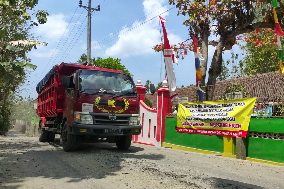 Truk melintas di jalan rusak depan SD Negeri 2 Wonorejo, Pedukuhan Gendol, Kalurahan Banyuroto, Kapanewon Nanggulan, Kabupaten Kulon Progo, Daerah Istimewa Yogyakarta. Debu jalan beterbangan saat truk lewat. Truk itu mengangkut batu ke penggilingan. Tampak pagar sekolah menggantung spanduk bertuliskan protes warga yang berharap suasana yang lebih sehat lewat perbaikan jalan.