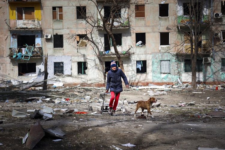 Seorang pria berjalan dengan anjingnya di dekat sebuah gedung apartemen yang rusak akibat penembakan di pinggiran Mariupol, Ukraina, di wilayah yang dikuasai pemerintah separatis Republik Rakyat Donetsk, pada Selasa, 29 Maret 2022. 