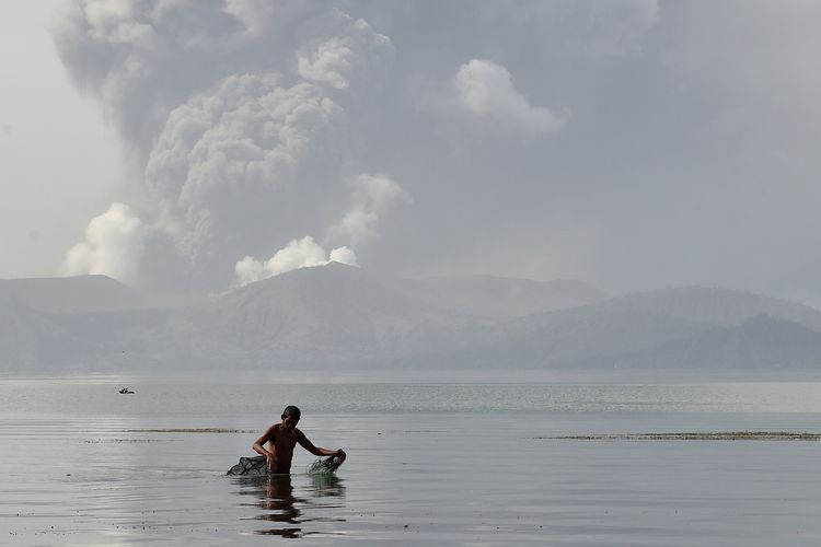 Seorang nelayan tengah menarik jalanya di perairan Danau Taal, dengan di belakangnya Gunung Taal memuntahkan abu pada 13 Januari 2020. Otoritas Filipina bersiaga dengan kemungkinan letusan dahsyat dari Gunung Taal.