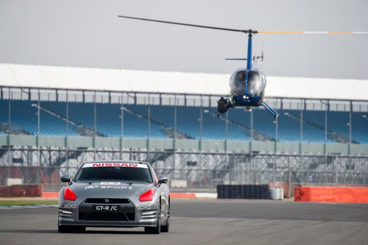 Mobil balap Nissan GT-R/C melaju di lintasan Silverstone sementara Jann Mardenborough mengendalikannya dengan gamepad PS4 dari helikopter.