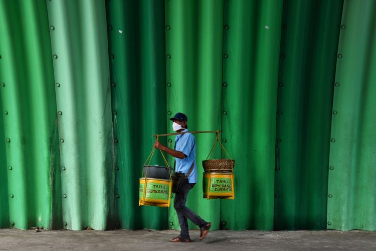 Terlihat salah satu pedagang mengenakan masker tengah berjalan memikul dagangannya di tengah pandemi virus corona di Bandung.