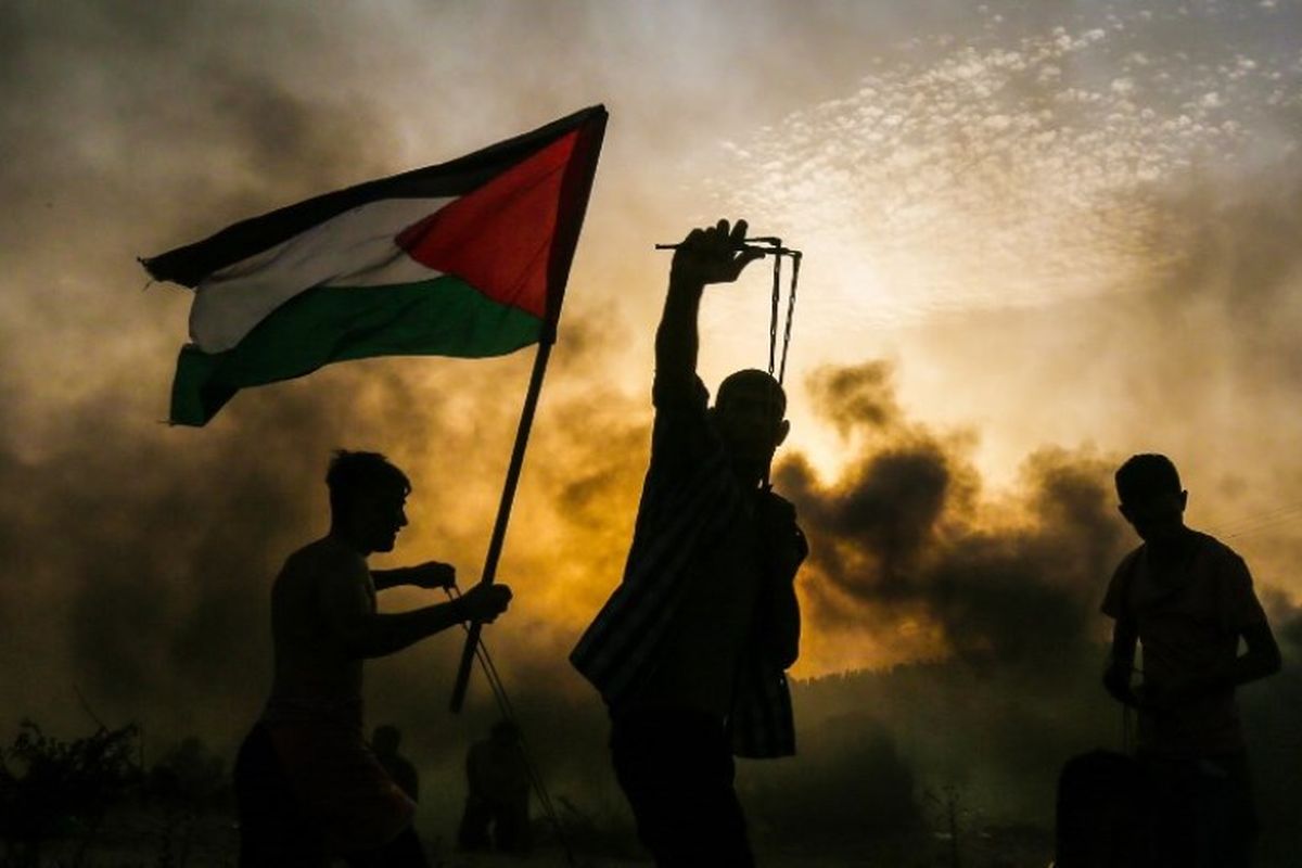 Seorang pengunjuk rasa Palestina memegang bendera saat pengunjuk rasa lainnya melemparkan batu ke arah pasukan Israel selama demonstrasi di pantai dekat perbatasan Israel, di Jalur Gaza utara, pada Senin (22/10/2018). (AFP/Mahmud Hams)