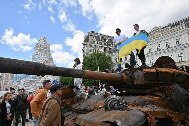 Warga Ukraina berpose di atas tank rusak Rusia sambil memegang bendera Ukraina pada pembukaan pameran perlengkapan rusak militer Rusia di pusat kota Kyiv, 28 Mei 2022.