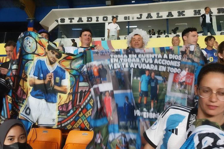 Legenda sepak bola Argentina, Diego Armando Maradona, hadir mendukung para junior penerusnya pada ajang Piala Dunia U17 2023. Spanduk tersebut terlihat ketika timnas U17 Argentina melawan Senegal pada matchday pertama Grup D di Stadion Si Jalak Harupat, Bandung, pada Sabtu (11/11/2023) malam WIB.