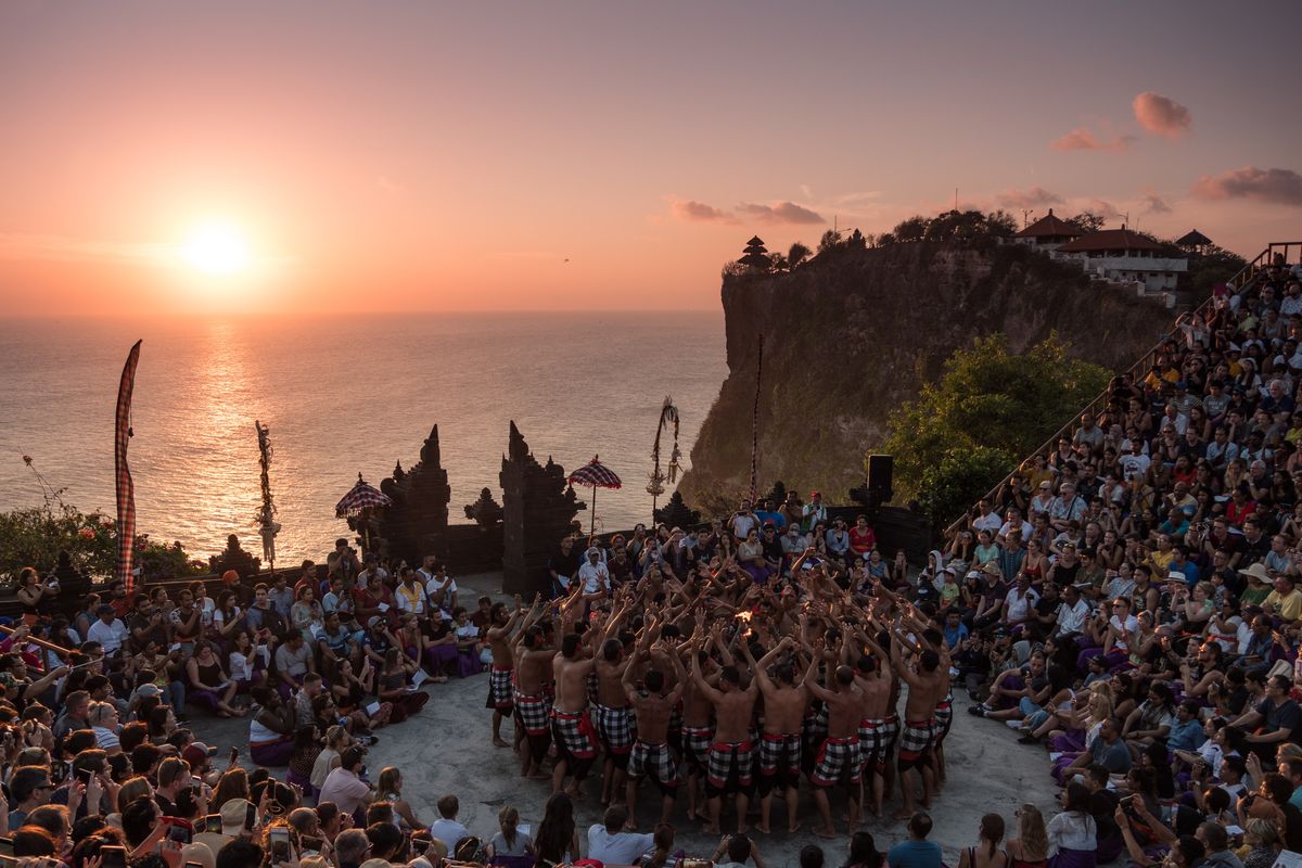Ilustrasi pertunjukkan tari kecak di kawasan Pura Luhur Uluwatu, Bali, dengan latar laut dan matahari terbenam.