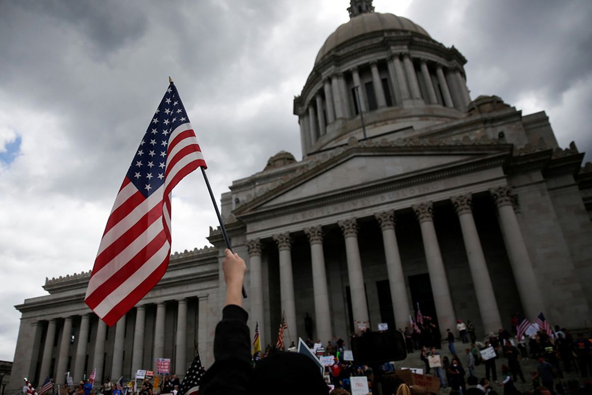 Seorang warga mengibarkan bendera Amerika saat protes atas perpanjangan perintah tetap berada di rumah untuk memperlambat penyebaran penyakit virus corona (Covid-19) di gedung Capitol di Olympia, Washington, Amerika Serikat, Minggu (19/4/2020). Berdasarkan data Johns Hopkins University, hingga Selasa (21/4/2020), AS masih menjadi negara dengan kasus Covid-19 tertinggi dunia mencapai 787.794 dengan korban meninggal 42.362.