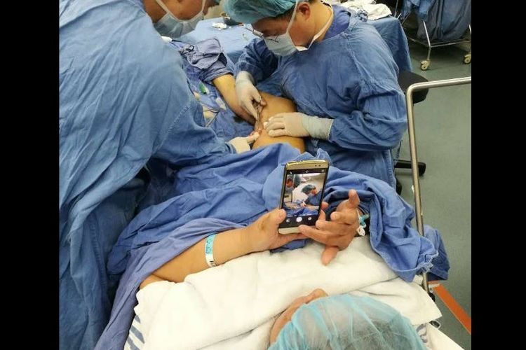 Seorang perempuan tetap memainkan telepon pintarnya saat menjalani pembedahan di kakinya.