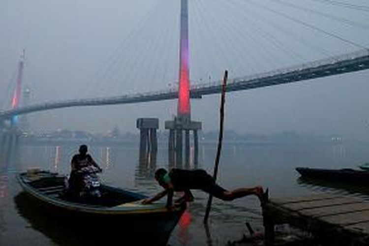 Pengendara sepeda motor memanfaatkan jasa tukang perahu untuk menyeberangi Sungai Batanghari di Jambi dengan latar belakang Jembatan Gentala Arasy yang diselimuti kabut asap, Senin (14/9/2015) sekitar pukul 18.00. Udara berasap bercampur abu kebakaran lahan semakin pekat dan mengganggu aktivitas serta kesehatan warga.