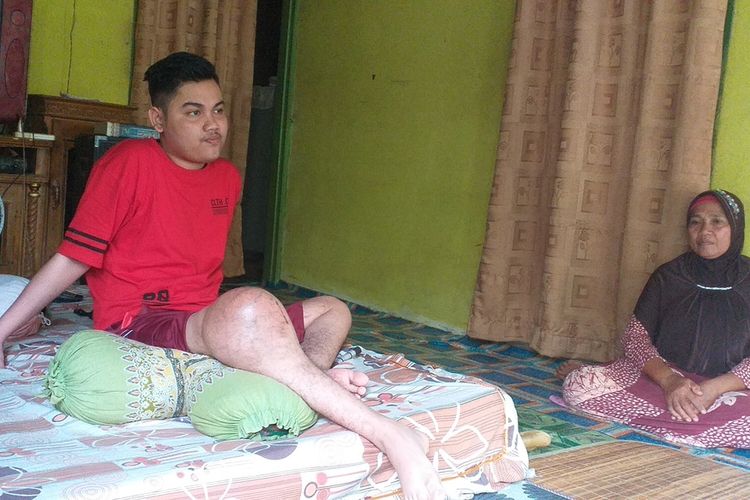 Marwan Alfidri (17), remaja yang mengidap tumor di kaki sebelah kakan sedang duduk di kasur ditemani ibunya, Siti Mariam, di ruang tengah rumahnya di Desa Pulau Jambu, Kecamatan Kampa, Kabupaten Kampar, Riau, Rabu (5/2/2020).