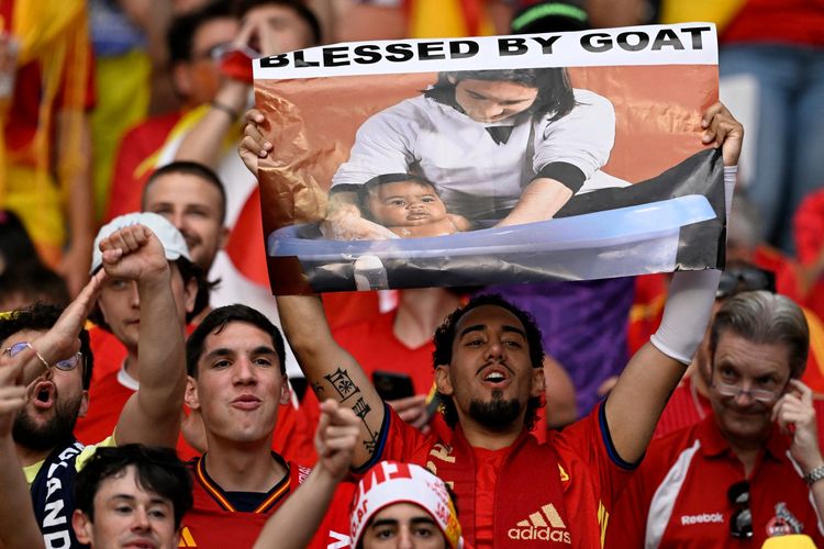 Seorang pendukung timnas Spanyol memegang spanduk dengan foto yang menunjukkan Lionel Messi dari Argentina berpose dengan penyerang muda Spanyol, Lamine Yamal, yang mengunjungi ruang ganti Barcelona sebagai bagian dari pemotretan kalender amal pada tahun 2007.