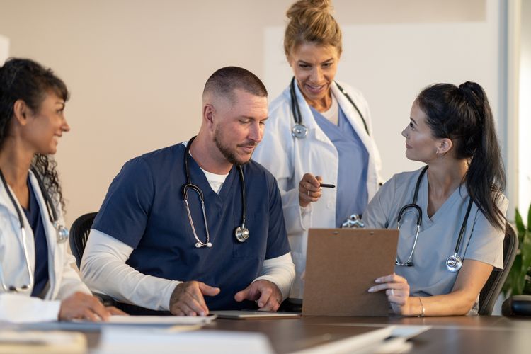 A small group of medical students sit at a desk together discussing patient cases. Their supervising doctor is standing behind them guiding them as they work through learning the career. They are each dressed professionally in medical scrubs and lab coats as they learn together.