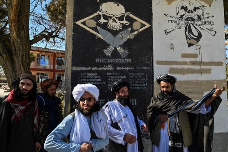 Dalam gambar yang diambil pada 15 November 2021, kepala budaya provinsi Taliban Mullah Habibullah Mujahid (kanan) berdiri dengan anggota Taliban di sebelah bagian dinding bekas pangkalan militer AS dengan nama tentara AS di Ghazni. 
