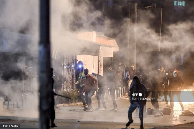 Para pemuda yang berbuat kerusuhan berdiri di tengah asap saat mereka memblokir jalan selama bentrokan dengan pasukan keamanan di pinggiran kota Ettadhamen di pinggiran barat laut ibu kota Tunisia, Tunis, pada 17 Januari 2021.
