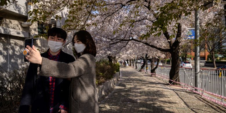Pejalan kaki yang mengenakan masker untuk mencegah virus corona, berfoto selfie di antara pohon bunga yang mekar di distrik Yeouido, Seoul, 5 April 2020.
