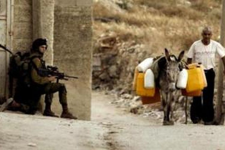 Foto ini diambil pada 26 Juni 2014, memperlihatkan seorang pria Palestina menuntun seekor keledai melewati para prajurit Israel yang sedang bersiaga dalam sebuah operasi militer di desa Awarta, Tepi Barat.