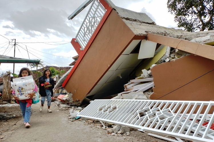 Seorang anak sambil membawa popok melintas di depan bangunan rumah yang ambruk di Desa Cibulakan, Cugenang, Cianjur, Jawa Barat, Rabu (23/11/2022).