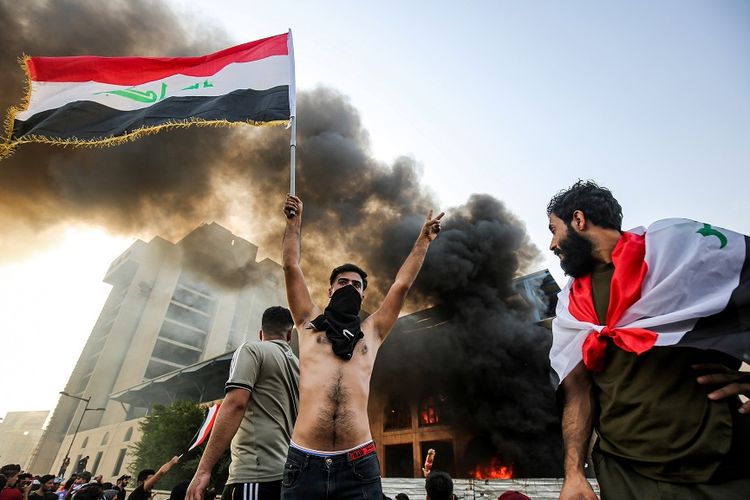 Pengunjuk rasa membawa bendera Irak di depan sebuah gedung terbakar di Baghdad, saat aksi unjuk rasa menentang korupsi di negara itu, Selasa (1/10/2019).