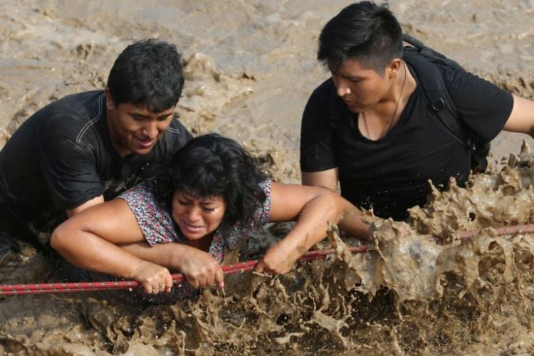 Dua anggota tim penyelamat menyeberangkan seorang perempuan di sebuah ruas jalan utama yang berubah menjadi sungai coklat dengan arus yang deras di Distrik Huachipa, Lima, ibu kota Peru, Jumat (17/3/2017)