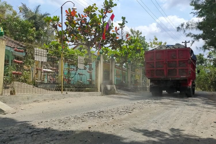 Truk melintas di jalan rusak depan SD Negeri 2 Wonorejo, Pedukuhan Gendol, Kalurahan Banyuroto, Kapanewon Nanggulan, Kabupaten Kulon Progo, Daerah Istimewa Yogyakarta. Debu jalan beterbangan saat truk lewat. Truk itu mengangkut batu ke penggilingan. Tampak pagar sekolah menggantung spanduk bertuliskan protes warga yang berharap suasana yang lebih sehat lewat perbaikan jalan.