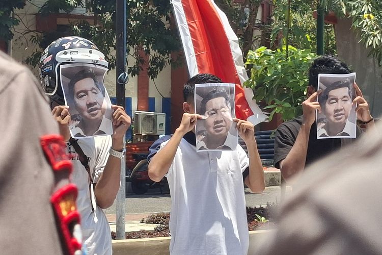 Pendemo menutupi wajahnya dengan gambar wajah Menteri Ara sebagai kritik. 