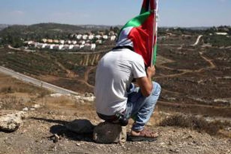 Seorang demonstranduduk sambil memegang bendera Palestina setelah aksi melawan perampasan tanah Palestina untuk memperluas pemukiman Yahudi Hallamish (latar belakang), Jumat (28/8/2015), di Desa Nabi Saleh, Tepi Barat, dekat Ramallah.