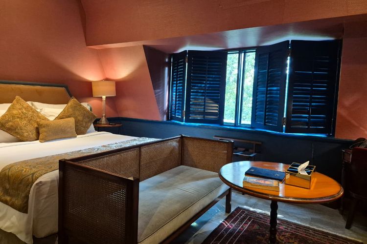 Warna dinding terracotta di The Mansard Room memberi kesan hangat sejak awal. Dipadukan dengan furnitur kayu gelap dan aksen biru tua, ruangan terasa tenang tanpa banyak elemen tambahan. Tempat tidur menjadi pusat perhatian, lengkap dengan headboard klasik dan bantal dekoratif bernuansa emas yang tetap terasa proporsional. 
