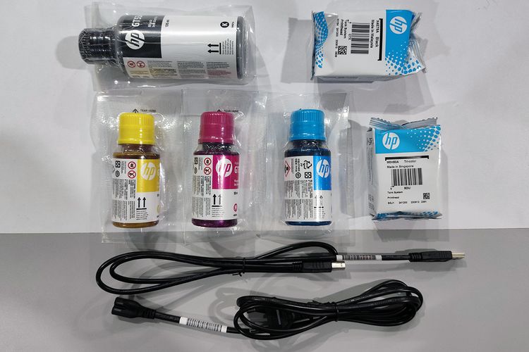 Serangkaian pelengkap dalam kemasan HP Smart Tank 580, mencakup botol tinta black, cyan, magenta, dan yellow, dua buah printhead, kabel USB A ke USB B, serta kabel daya. 