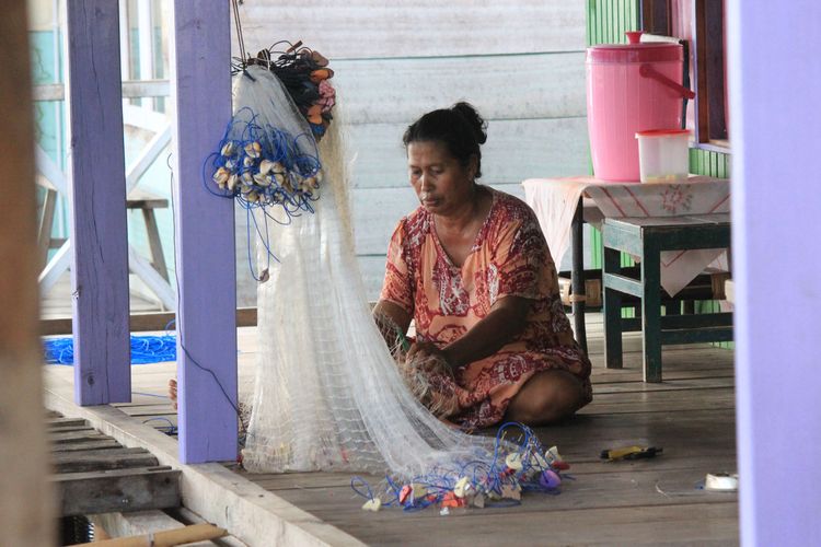Seorang wanita suku bajau tengah memperbaiki jaring penangkap ikan di teras rumah. wanita bajau dituntut untuk melakukan banyak hal pekerjaan rumah tanga hingga mencari ikan.
