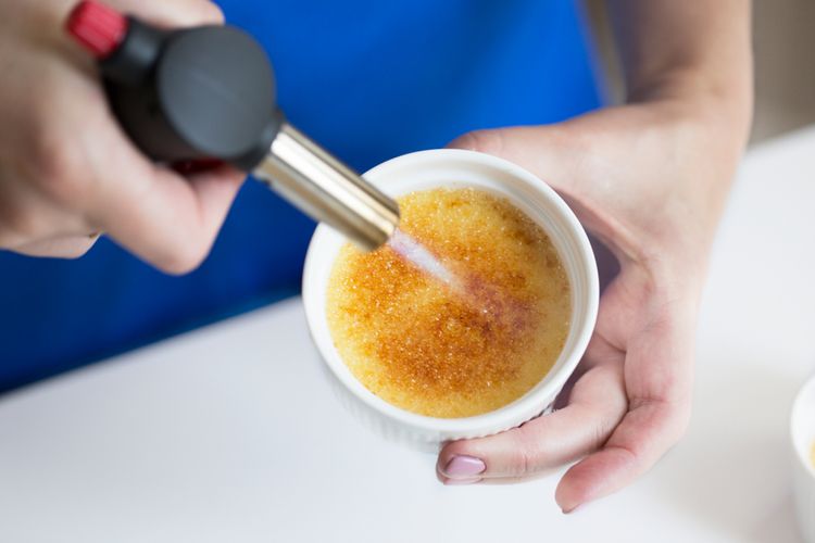 Ilustrasi karamel creme brulee sedang digosongkan menggunakan torch. 