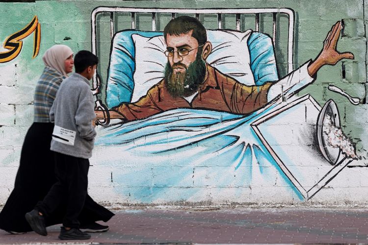Seorang wanita Palestina dan putranya berjalan melewati mural yang menggambarkan Khader Adnan, seorang pemogok makan Palestina dan tokoh terkemuka dalam kelompok Jihad Islam di Tepi Barat yang diduduki, di Kota Gaza pada 2 Mei 2023. Khader Adnan memulai mogok makan pada 5 Februari 23 dan dinyatakan meninggal pada 2 Mei 2023. 